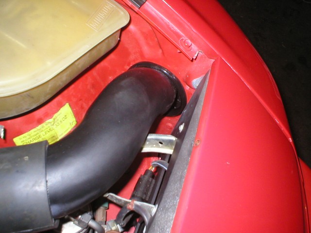 The pipe exits the engine bay through the stock air intake hole after passing over the headlight stop adjustment arm and linkage.